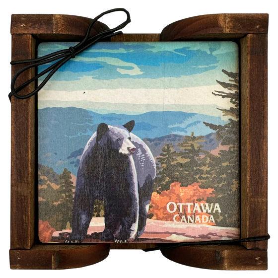 A square wooden coaster displayed in an attractive wooden holder. The coaster depicts  a black bear standing on a rock against a background of forest, mountains, and blue sky. In the bottom right, the coaster reads "Ottawa Canada"