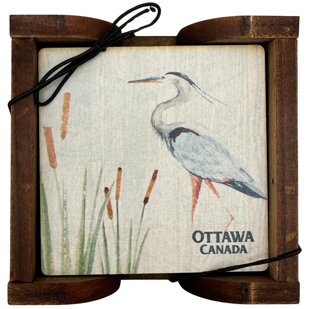 A square wooden coaster contained in an attractive holder. The coaster shows a majestic blue heron standing in reeds against a white background and "Ottawa, Canada" written on the bottom.