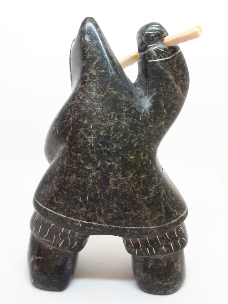 Back side view of soapstone carving of Inuit hunter figure. The etchings marked in the stone gives the figure the appearance of wearing a large hooded jacket and boots. Figure has right arm upstretched with hunting tool in hand. The tool is a carved white bone with jagged end.