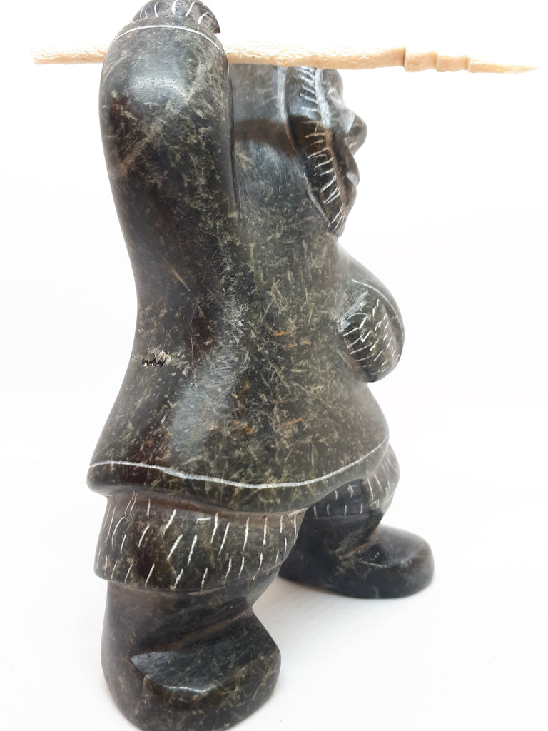 Side view of soapstone carving of Inuit hunter figure. The etchings marked in the stone gives the figure the appearance of wearing a large hooded jacket and boots. Figure has right arm upstretched with hunting tool in hand. The tool is a carved white bone with jagged end.