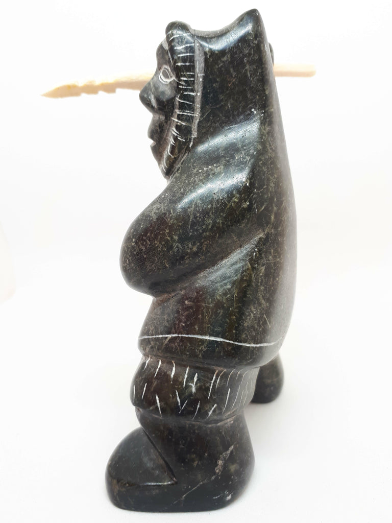 Side view of soapstone carving of Inuit hunter figure. The etchings marked in the stone gives the figure the appearance of wearing a large hooded jacket and boots. Figure has right arm upstretched with hunting tool in hand. The tool is a carved white bone with jagged end.