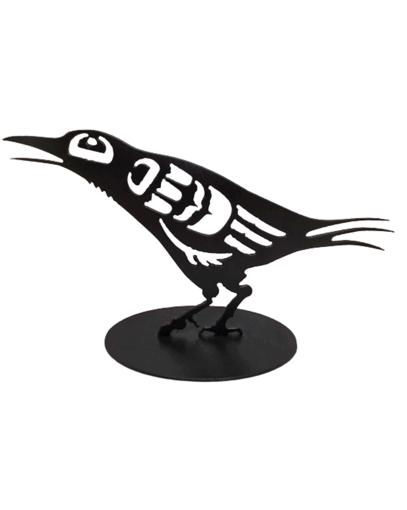 This metal sculpture shows the matte black silhouette of a baby crow drawn in Coastal Salish style. It is leaning forward with its beak slightly open. The slim tail, beak, and feather forms on this crow give it a dainty appearance.