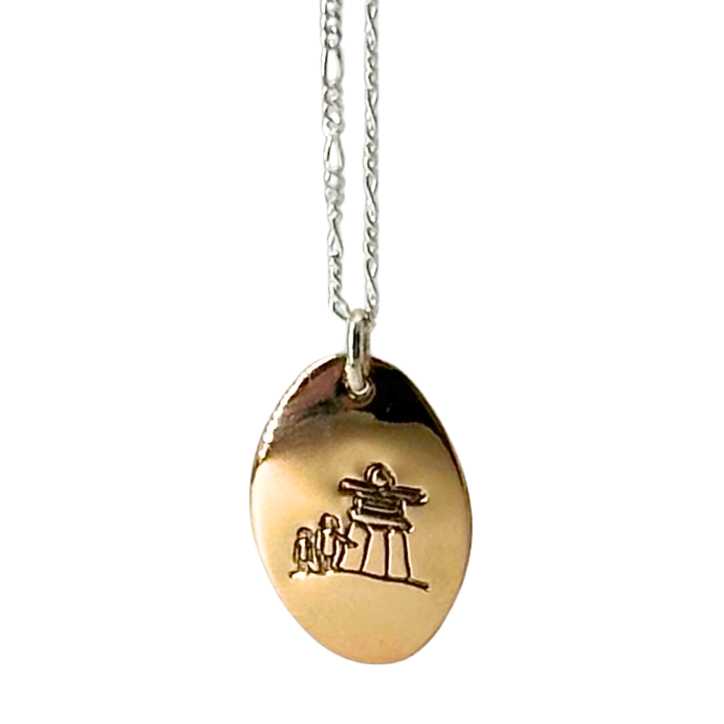 A necklace made of 14 karat gold fill and sterling silver on a gold chain. The base is an oval of bright gold with a large inukshuk with two smaller human figures etched into the centre.