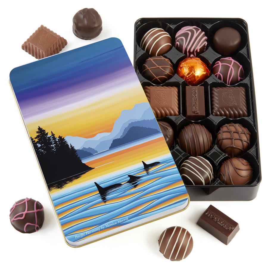 Delicious assorted chocolates in a stylish tin. On the cover of the tin, a beautiful sunset lights up a nature scene with mountains, trees and ocean. A family of three orca whales swim in the foreground.