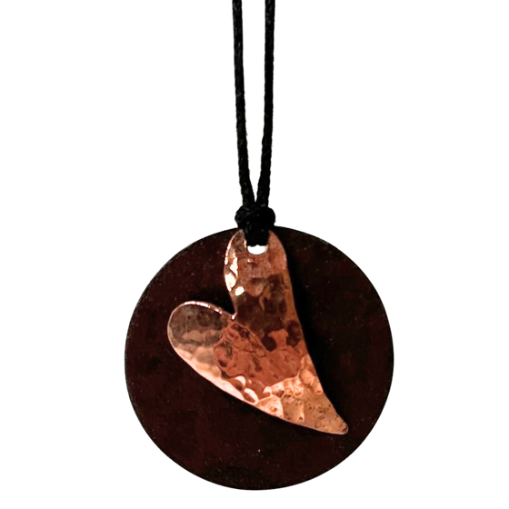 A circular heart necklace mounted on leather cord. The pendant is made of two layers: antique upcycled tin cut into a circle forms the base, and then a hammered copper heart is overlaid to create stunning contrast.