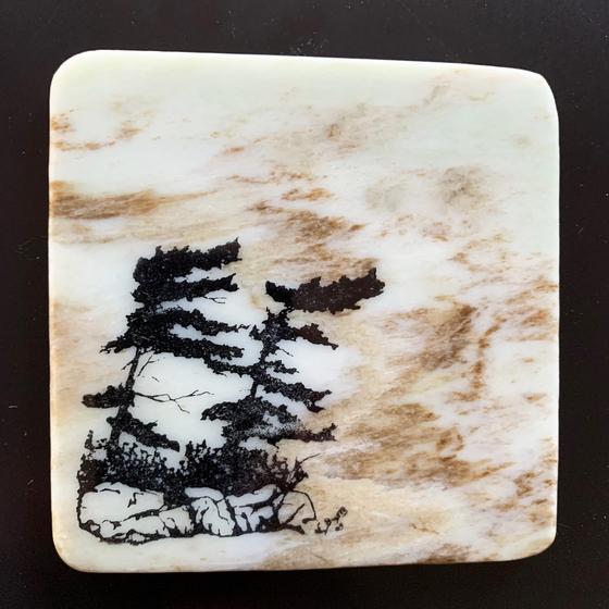 This coaster features the image of two pine trees on a rocky ledge with grass growing underneath. The image is on a piece of canadian shield marble with mineral lines running through in unique colours, lines, and patterns. The coaster is finished with a clear coat, giving it a shiny finish. 