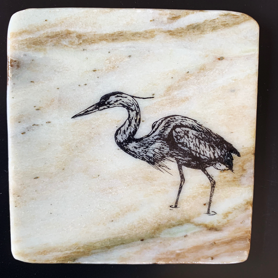 This coaster features the image of the side view of a heron as it walks through water. The image is on a piece of canadian shield marble with mineral lines running through in unique colours, lines, and patterns. The coaster is finished with a clear coat, giving it a shiny finish. 