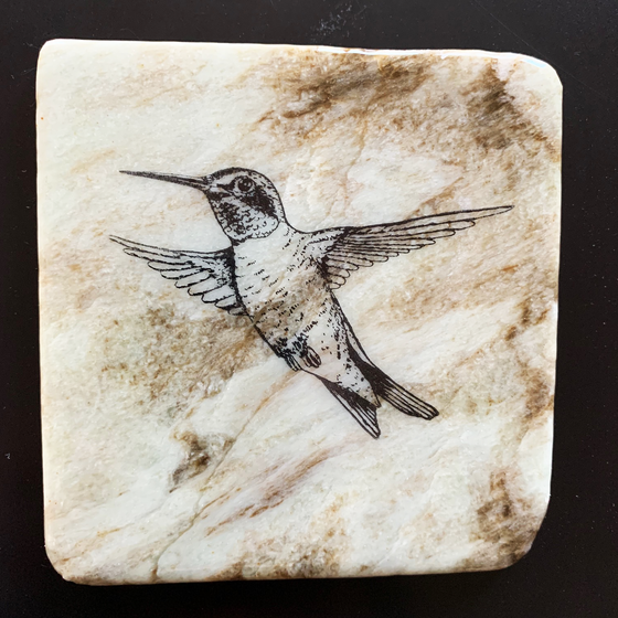 This coaster features the image of a hummingbird with its wings out in mid-flight. The image is on a piece of canadian shield marble with mineral lines running through in unique colours, lines, and patterns. The coaster is finished with a clear coat, giving it a shiny finish. 