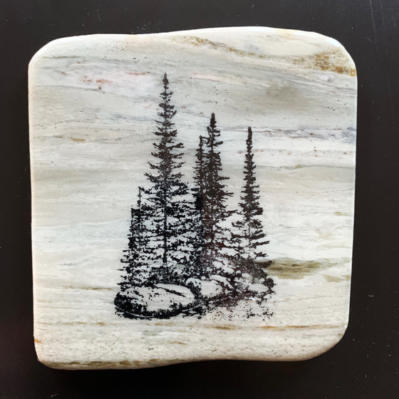 This coaster features the image of a small group of trees on a rocky ledge with grass growing around them. The image is on a piece of canadian shield marble with mineral lines running through in unique colours, lines, and patterns. The coaster is finished with a clear coat, giving it a shiny finish. 